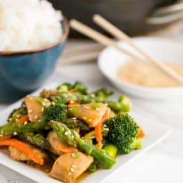 A plate of orange chicken and vegetable stir fry with a bowl of rice behind it